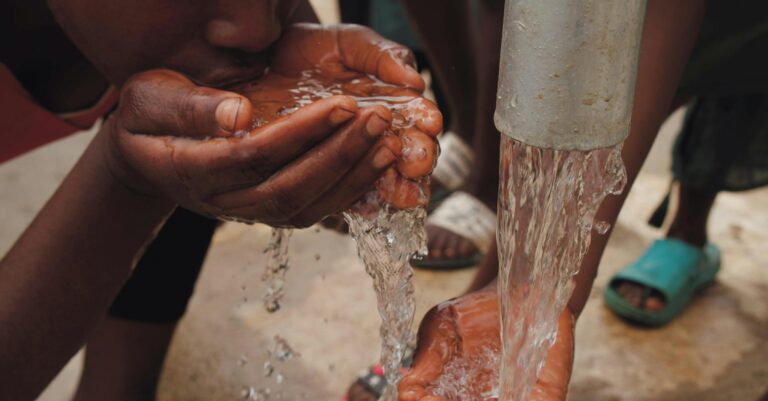 Close-up of hands and face drinking fresh water from an outdoor tap, emphasizing refreshment and necessity.